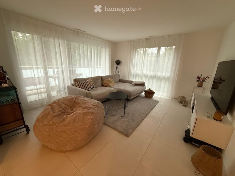 Modern living room with large windows, white curtains, grey sofa, coffee table, bean bag chair, television, and decorative items.