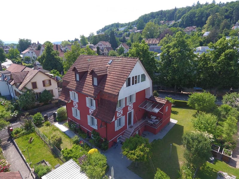 A two-story red house with a brown tiled roof, white trim, and multiple windows, surrounded by a well-maintained garden and trees.