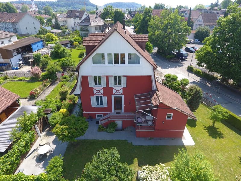A two-story red house with white trim and shutters, surrounded by a well-maintained garden and outdoor seating area.