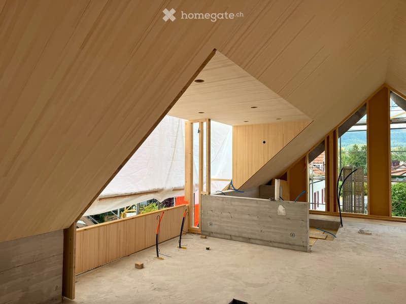 An attic space with wooden walls and ceiling, exposed beams, and large windows offering a view of the surrounding area. The floor is unfinished concrete, and the room is currently unfurnished.