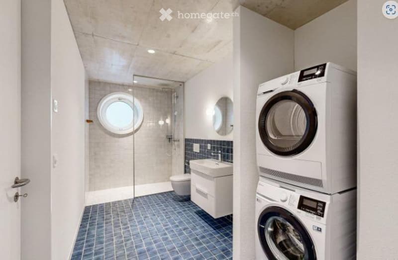 Modern bathroom with blue tiled floor, white walls, a glass-enclosed shower, a sink, a toilet, and a stacked washer/dryer unit.