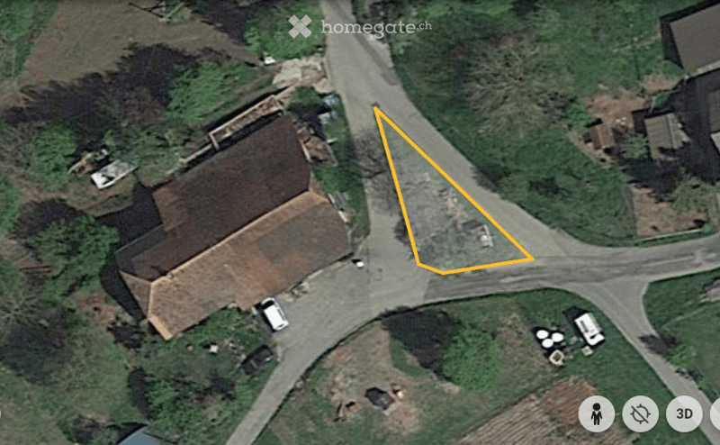 Aerial view of a rural building with a pitched roof, surrounded by greenery and a road. The building appears to be a house or small structure with a visible entrance and a small parking area nearby.