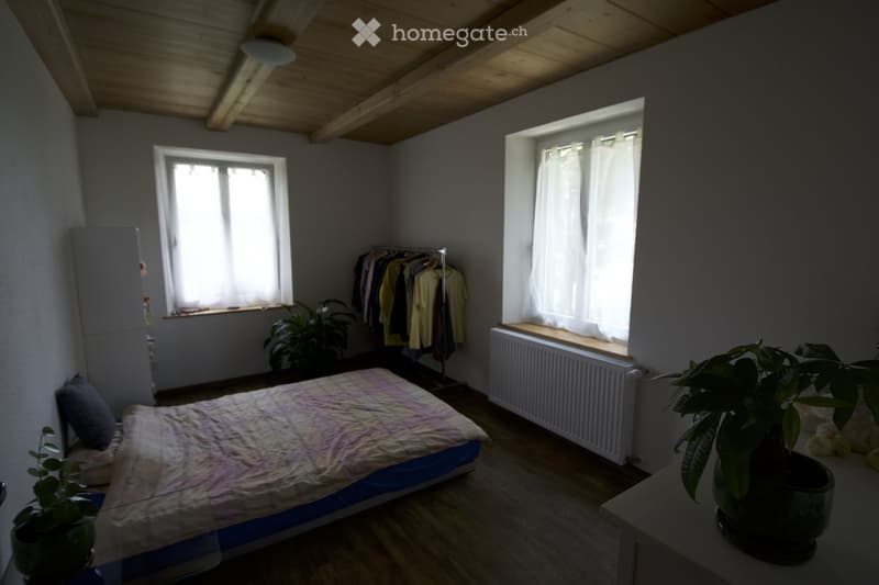Bedroom with wooden floor, slanted wooden ceiling, two windows with white curtains, bed with patterned bedspread, small plant on nightstand, radiator under window, wardrobe, and clothing rack with hanging clothes.