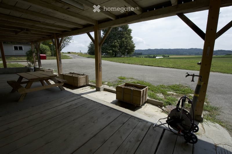 Wooden deck with a picnic table, two wooden planters, a faucet, and a cable reel, overlooking a rural landscape with fields and a road.