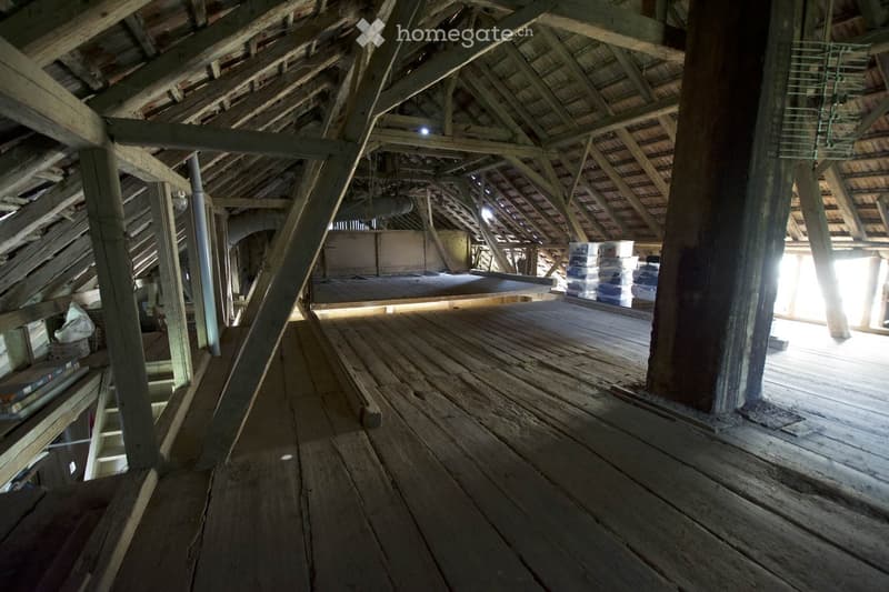 An attic space with exposed wooden beams and a wooden floor, featuring a partially constructed platform and some stacked materials in the background.