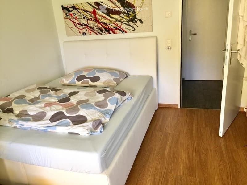 A bed with white mattress, wooden floor, white headboard, bedspread with abstract patterns, pillow, entrance door