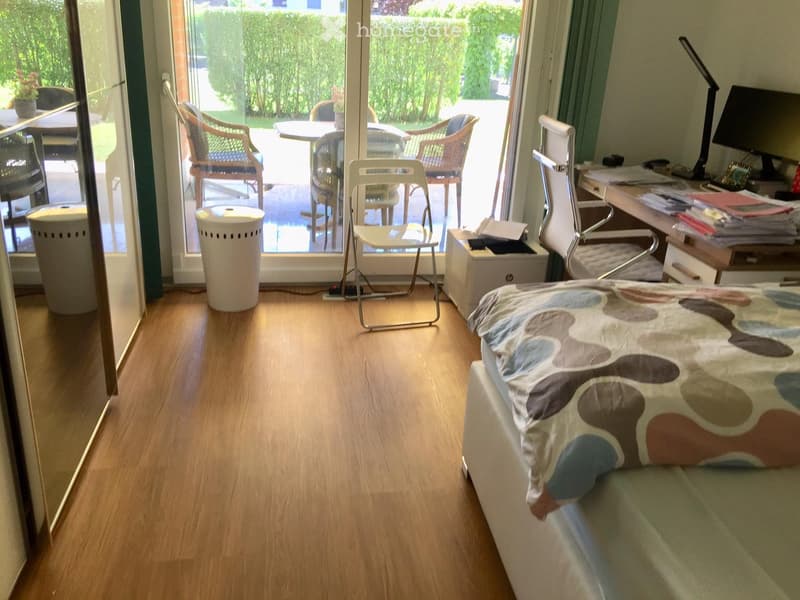 A room with wooden floor, bed, desk, chairs, sliding glass door, balcony, wastebaskets, monitor, lamp, papers, potted plants, and a table with flowers.