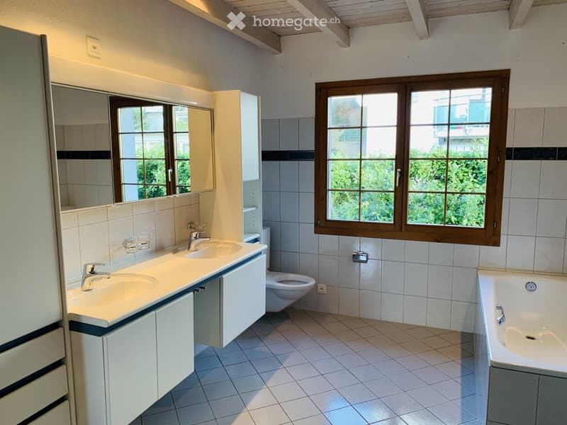 Modern bathroom with white tiled walls and floor, double sink vanity with mirrors, toilet, bathtub, and large window with wooden frame.