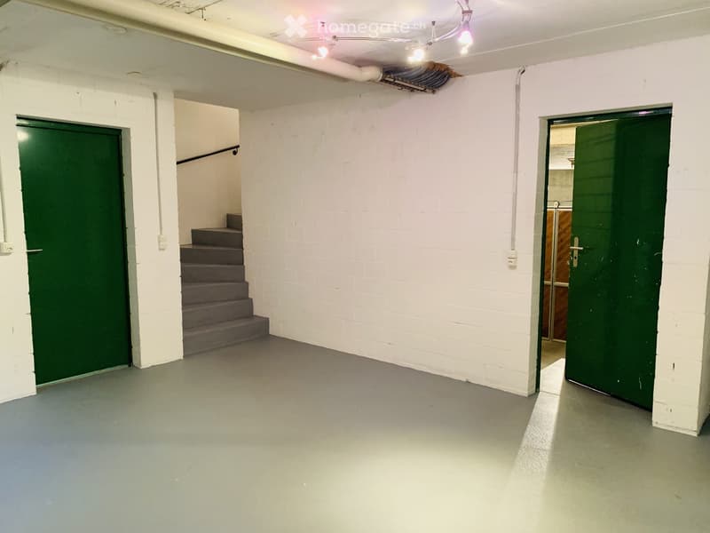 Empty basement room with white brick walls, concrete floor, green doors, and a staircase leading upwards.