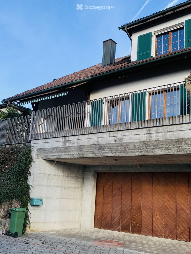 Two-story house with a tiled roof, green shutters, a balcony with metal railing, and a wooden garage door.