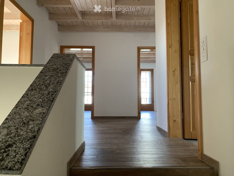 Spacious hallway with wooden flooring, white walls, and wooden doors leading to other rooms. Features a staircase with a granite railing and exposed wooden beams on the ceiling.