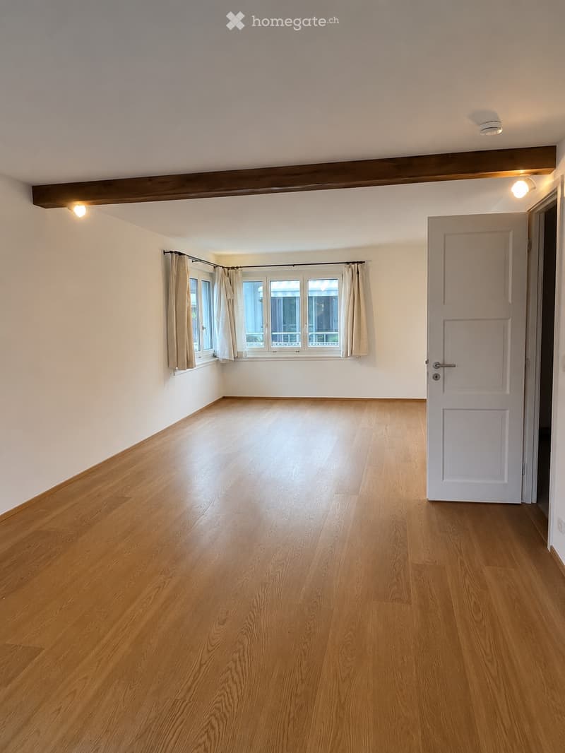 Empty room with wooden flooring, white walls, and large windows with beige curtains. The room has a white door and recessed lighting.
