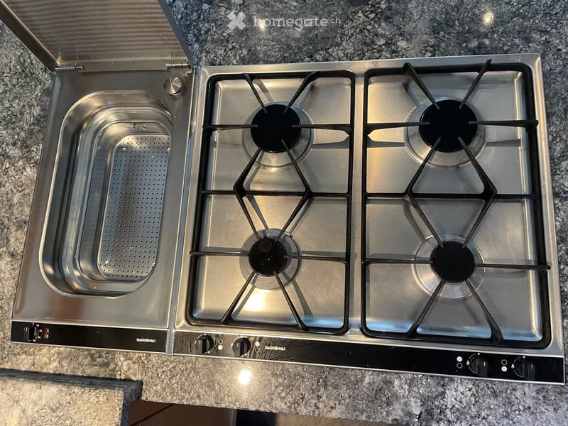 Modern kitchen with a stainless steel gas stove featuring four burners and a sink, placed on a granite countertop.
