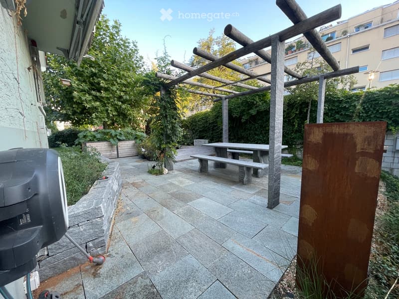 Outdoor patio with stone flooring, wooden pergola, stone benches, and a dining table surrounded by greenery and plants.