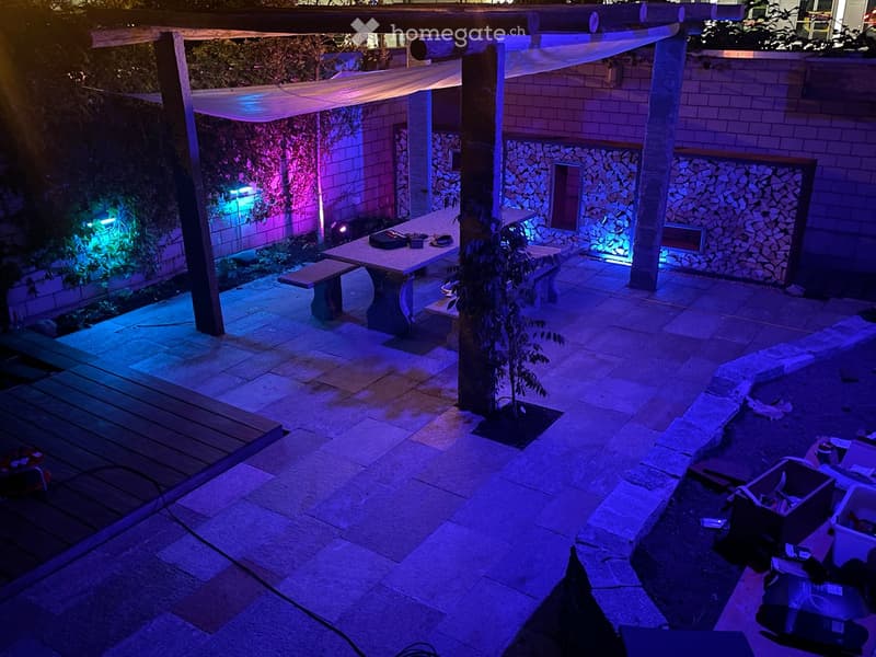 An outdoor patio area with a stone floor, a wooden pergola, a table with benches, and a small decorative fireplace surrounded by stacked stones.
