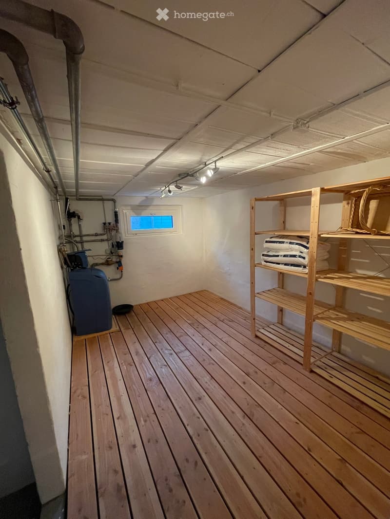 Empty room with wooden flooring, white walls, exposed pipes, a small window, and a wooden shelf unit with storage space.