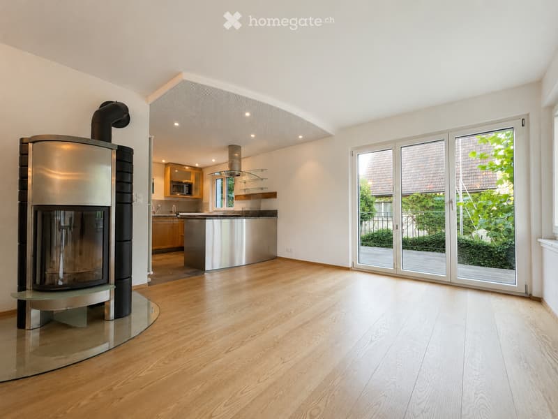 Modern living space with a central fireplace, wooden flooring, large sliding glass doors leading to a garden, and an open kitchen area with cabinets and a kitchen island.