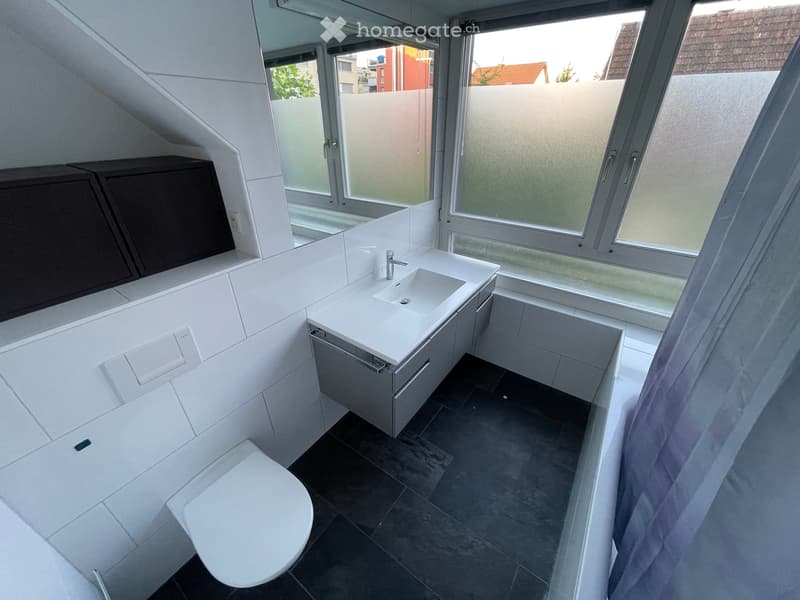 Modern bathroom with white tiles, a sink with a cabinet, a toilet, and large windows offering a view of the outside.