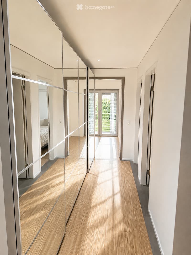 Modern hallway with light wooden flooring, white walls, and large mirrored panels on one side. The hallway leads to a glass door opening to a garden area.