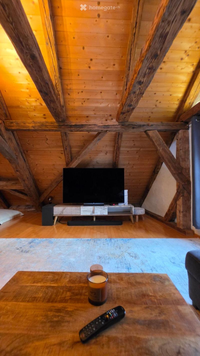 Cozy living room with wooden beams, television on a wooden stand, wooden coffee table with a candle and remote control, and a small stool.