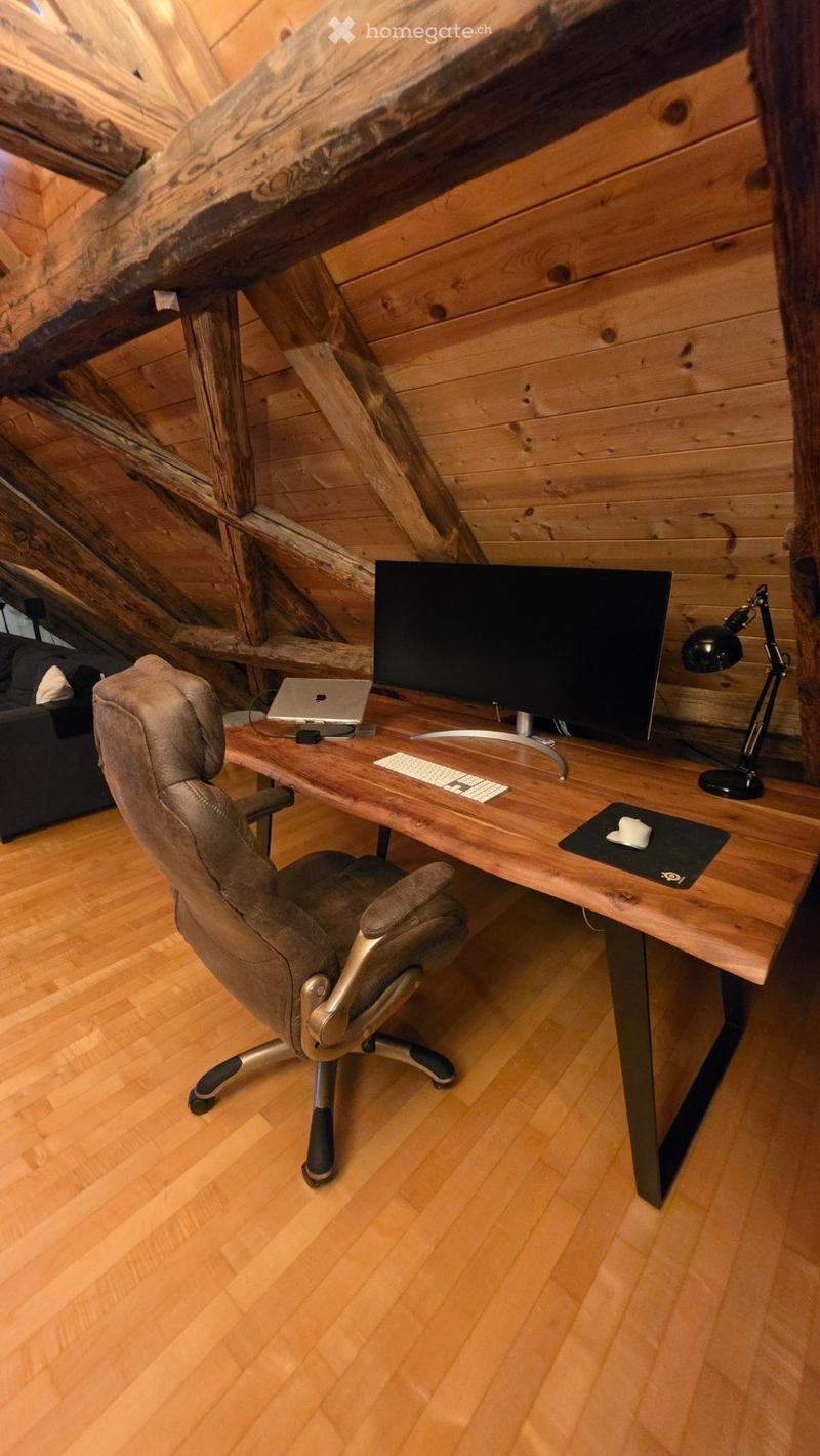 A modern, wooden-floored room with a large wooden desk, ergonomic chair, laptop, monitor, keyboard, mouse, and a desk lamp. Exposed wooden beams and a slanted ceiling add a rustic touch.