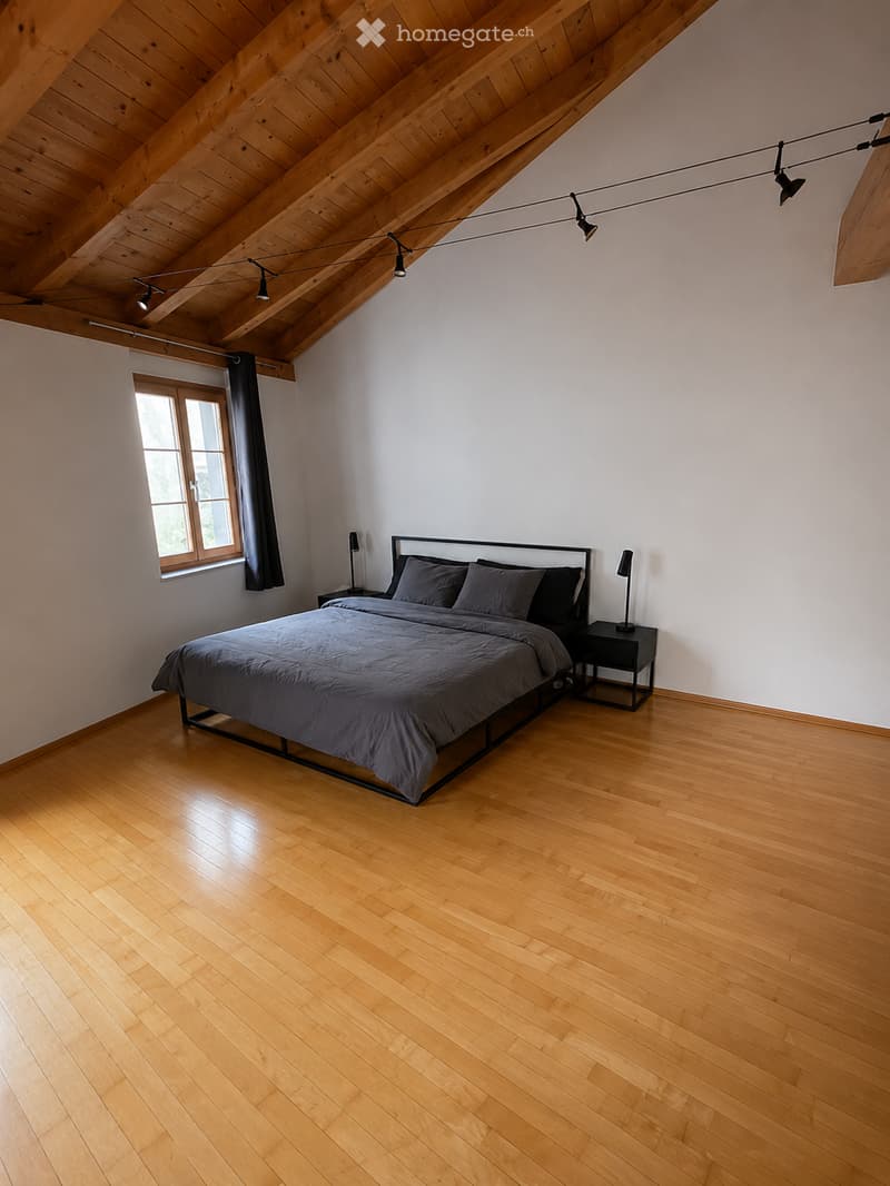 Modern bedroom with wooden floor, slanted wooden ceiling, large window with dark curtains, bed with dark bedding, nightstand with lamp.