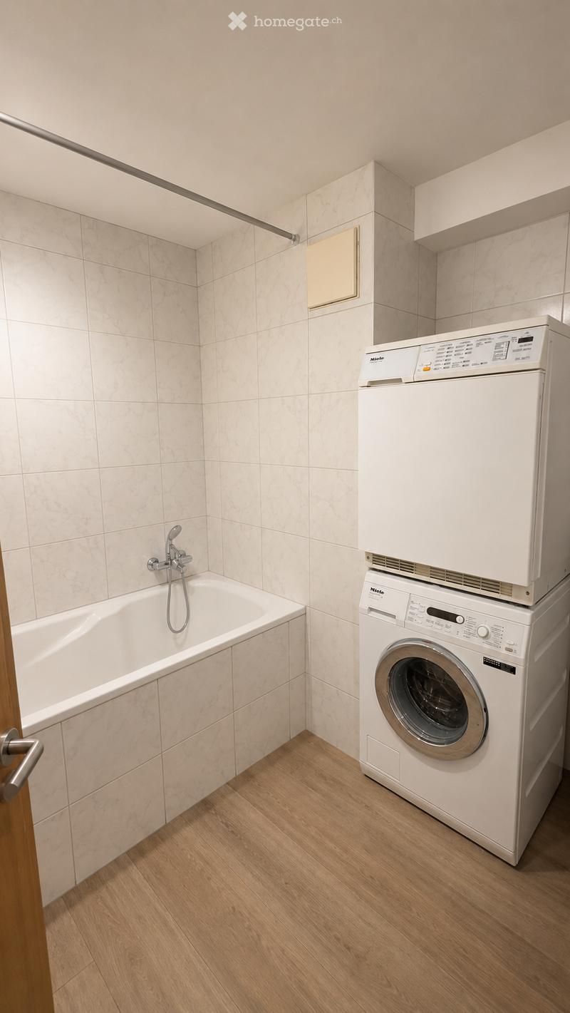 Bathroom with a bathtub, washing machine, and dryer. Light-colored tiles and wooden flooring.
