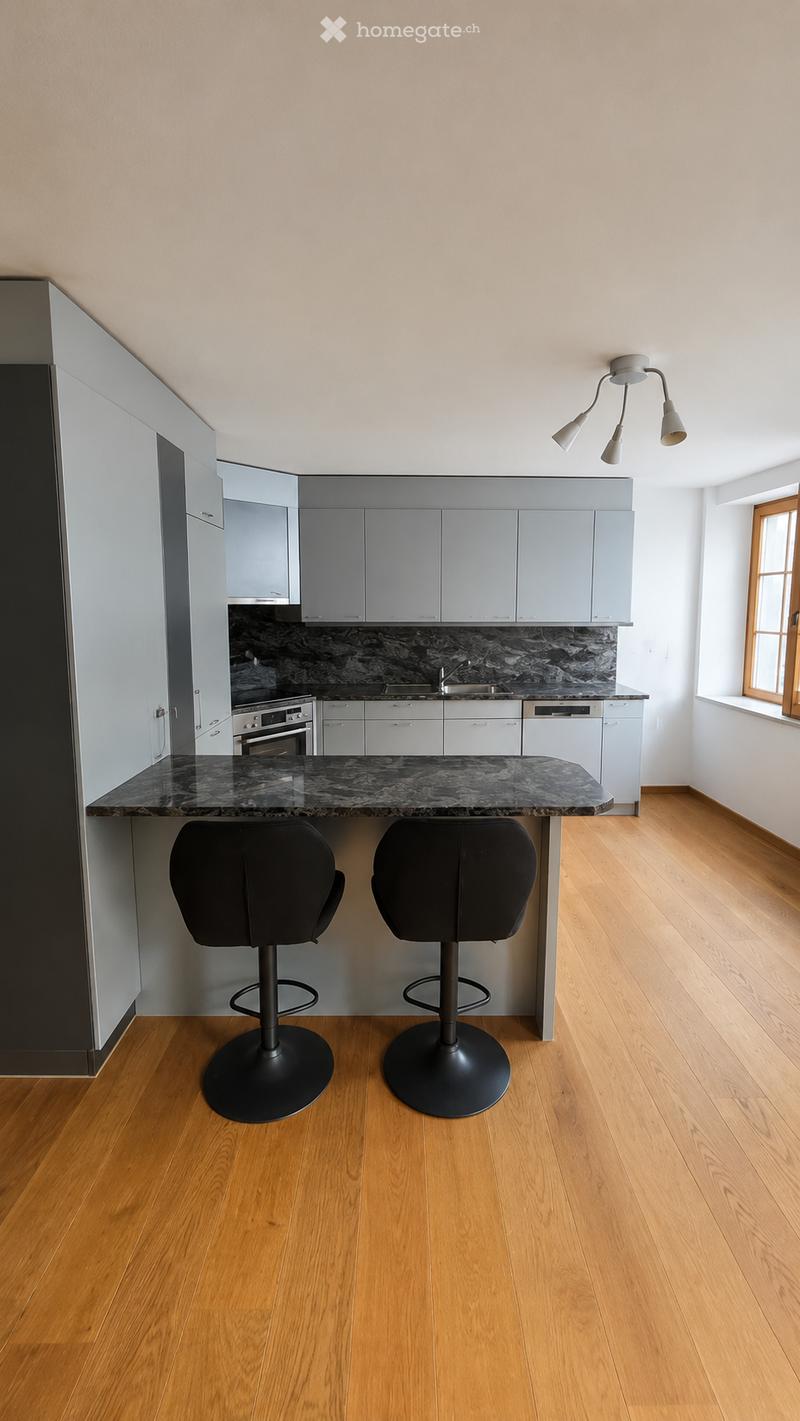 Modern kitchen with light grey cabinets, marble countertop, two black bar stools, and built-in appliances including an oven, sink, and dishwasher.