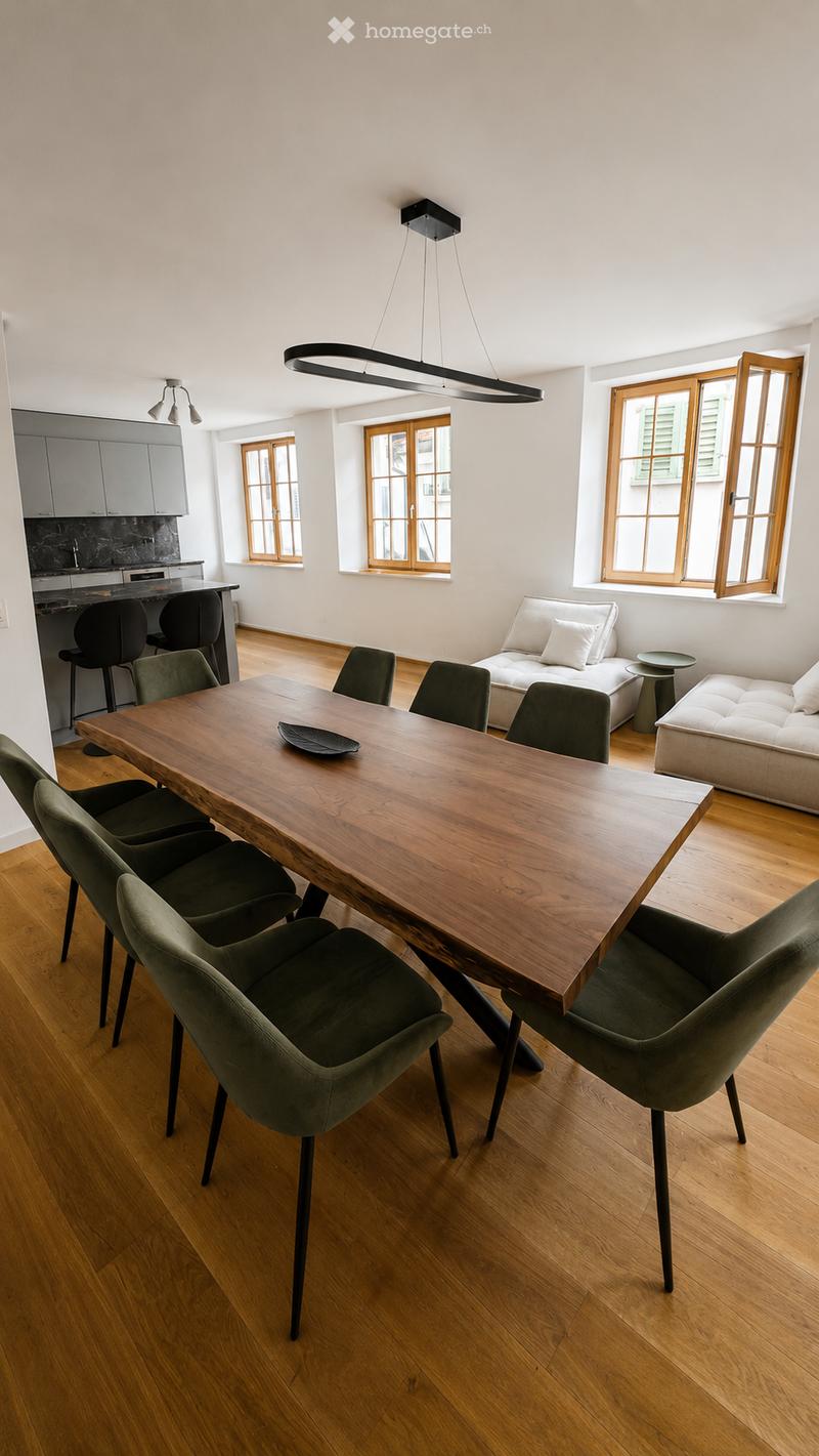 Open-plan living and dining area with wooden flooring, large windows, a long wooden dining table surrounded by green chairs, a white sofa, and a modern kitchen area with grey cabinets and a black countertop.