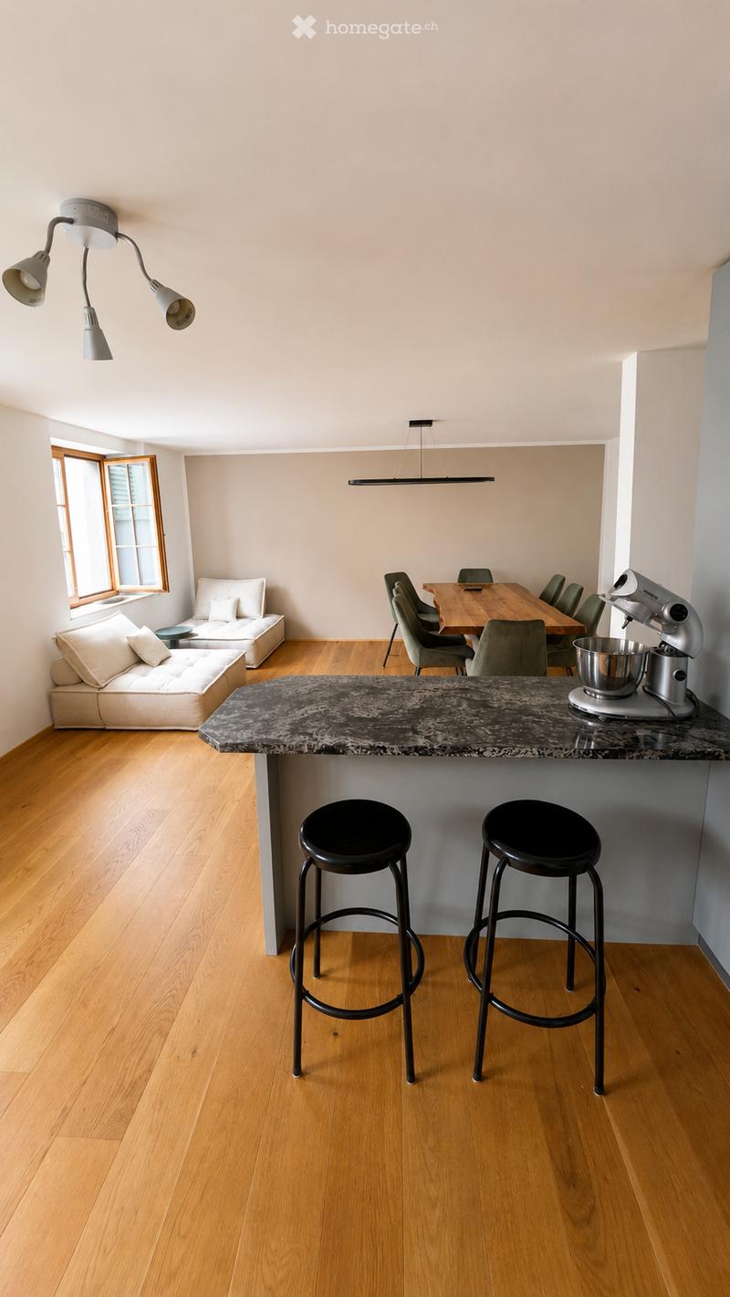 Open-plan living and dining area with light wood flooring, a white sofa, a kitchen island with a marble countertop, a stand mixer, and a dining table with chairs.
