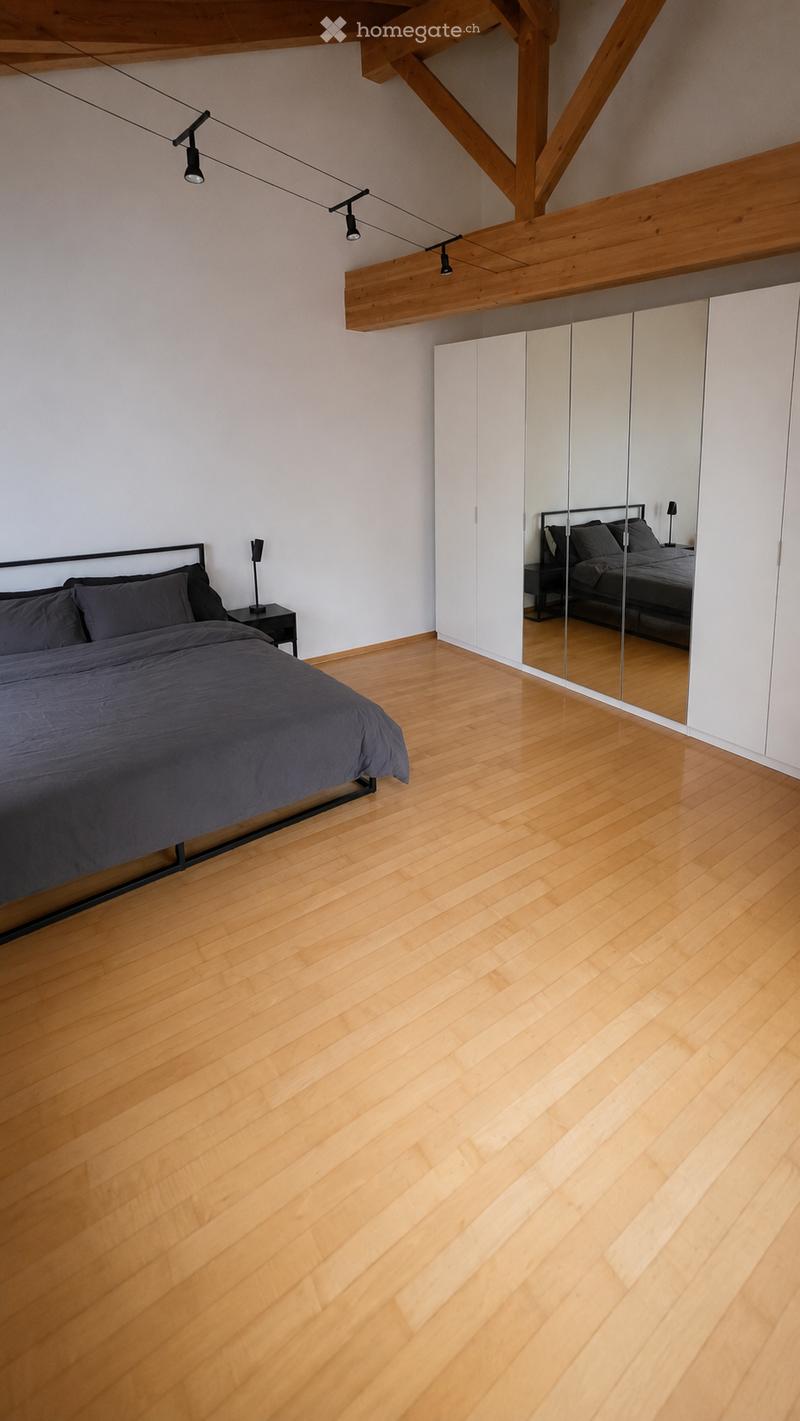 Modern bedroom with wooden flooring, a large bed with dark bedding, a nightstand with a lamp, and a mirrored wardrobe with built-in storage.