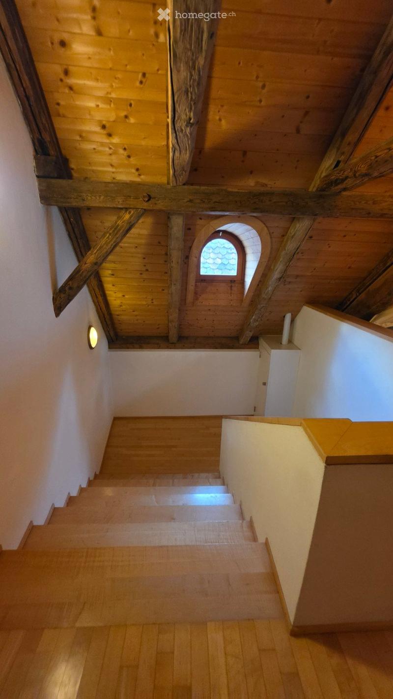 Wooden staircase with exposed wooden beams and a small arched window at the top, leading to an attic space with wooden ceilings and walls.
