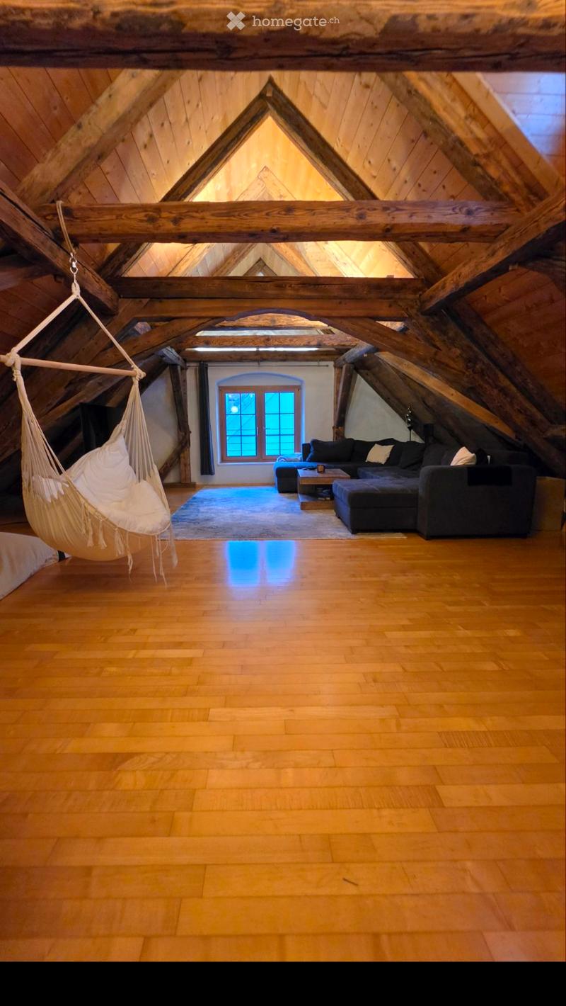 Attic living space with wooden beams, exposed wooden ceiling, hardwood floor, large window, hammock, sofa, coffee table, and ottoman.