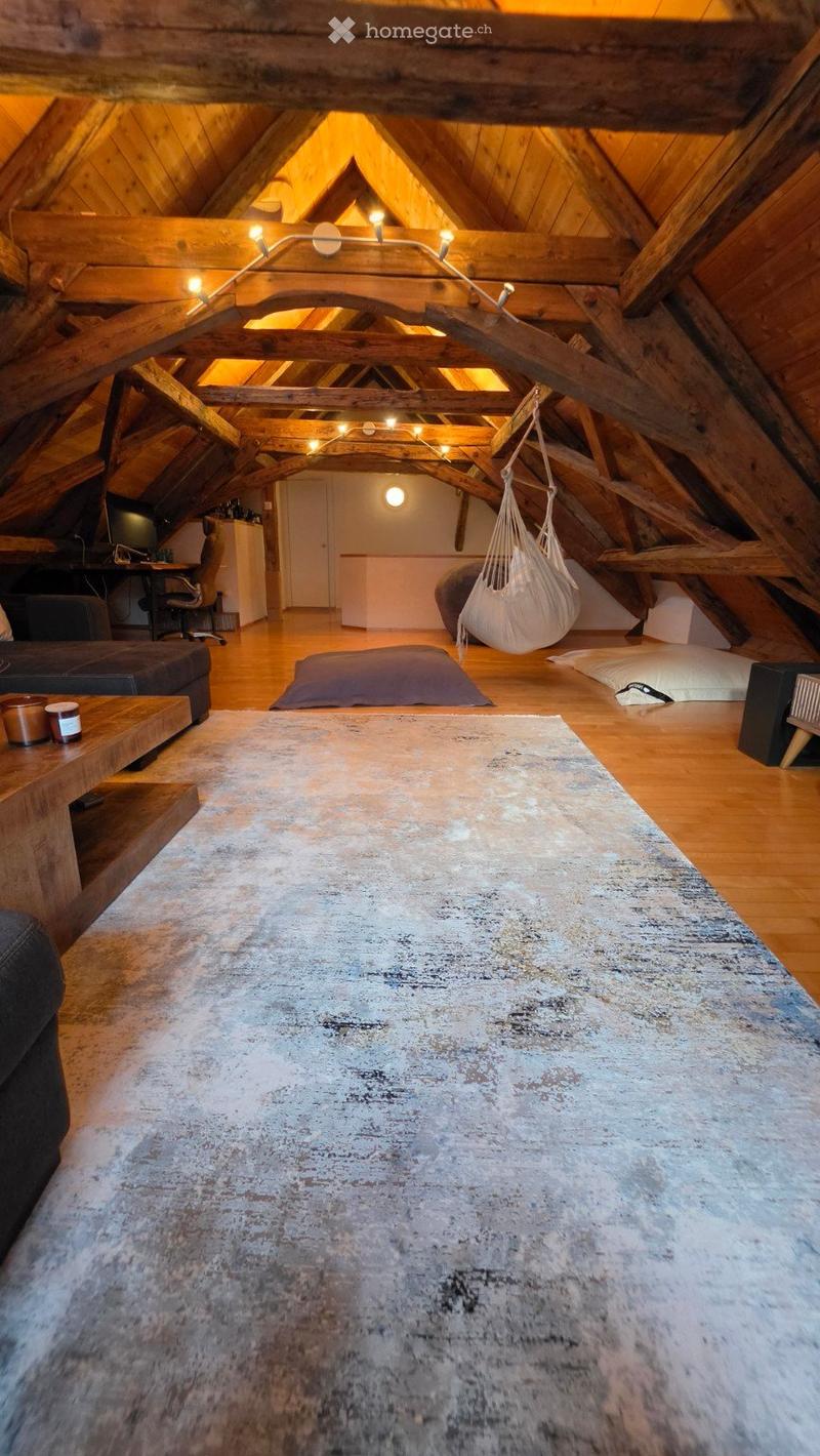 Loft-style living space with exposed wooden beams, a large area rug, a sofa, coffee table, hanging wicker chair, and a desk area.