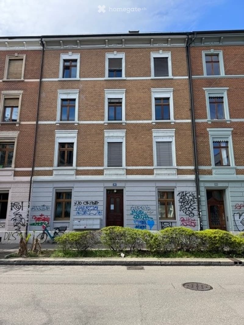 Four-story brick building with multiple windows, graffiti on the lower facade, and a small garden area in front.