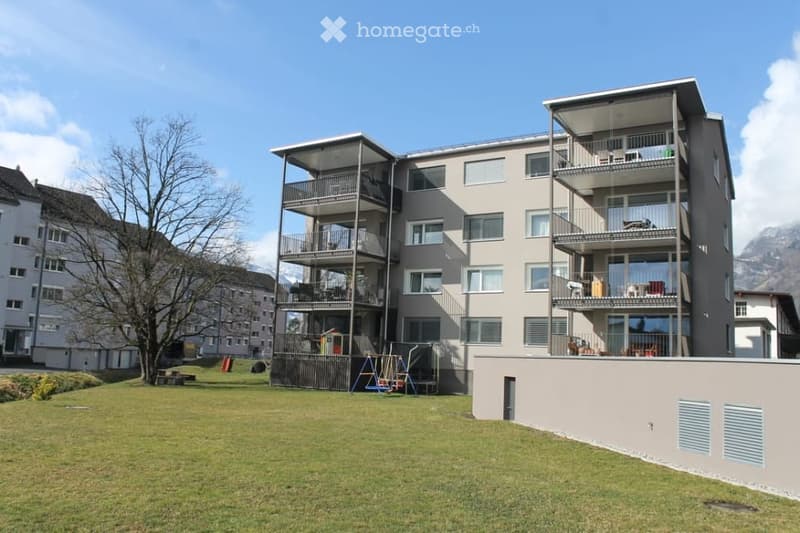 Modern multi-story apartment building with multiple balconies, large windows, and a view of mountains in the background. The building is surrounded by a grassy area with playground equipment.