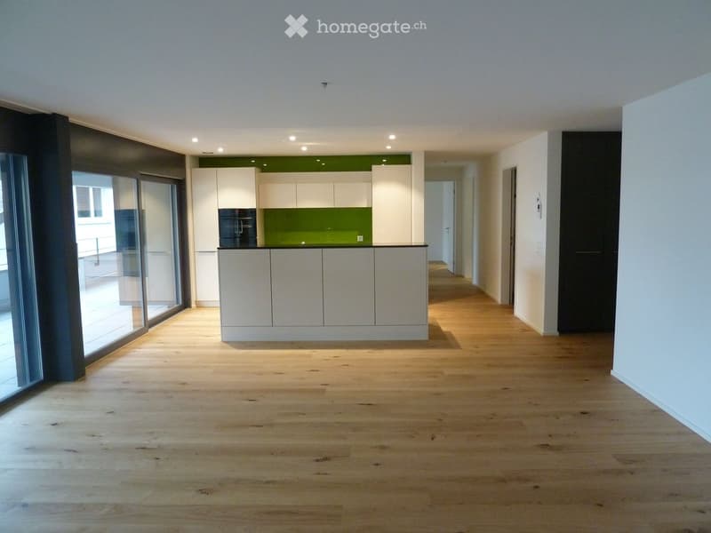 Empty room with light wooden flooring, white walls, and a small kitchen area with white cabinets and a green backsplash visible in the background.
