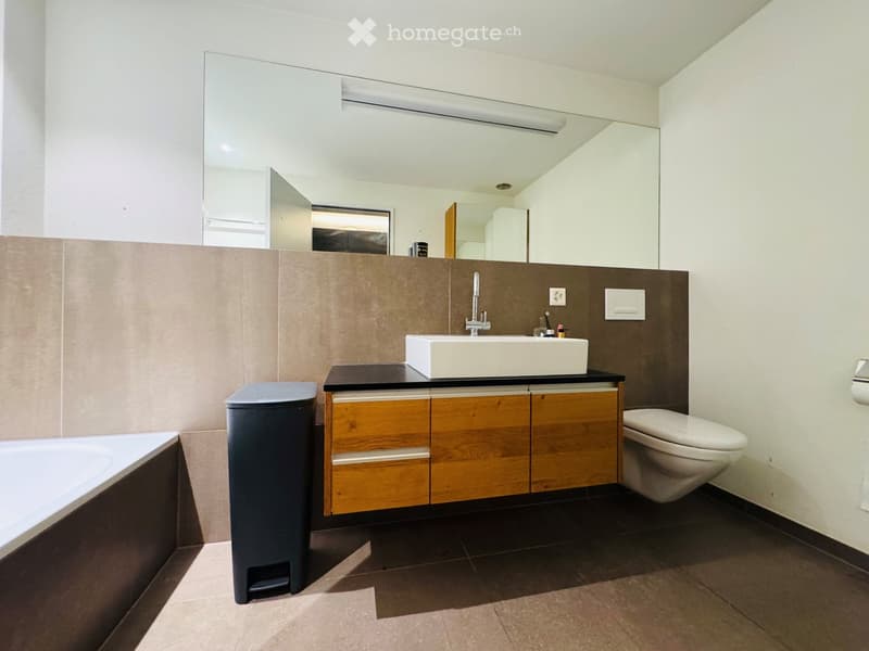 Modern bathroom with a white sink, wooden cabinet, large mirror, toilet, bathtub, and a dark trash bin.