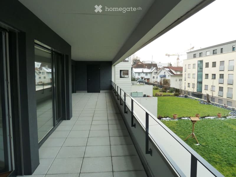 Modern balcony with tiled floor, glass railing, and view of a landscaped garden with grass, potted plants, and a bird feeder.