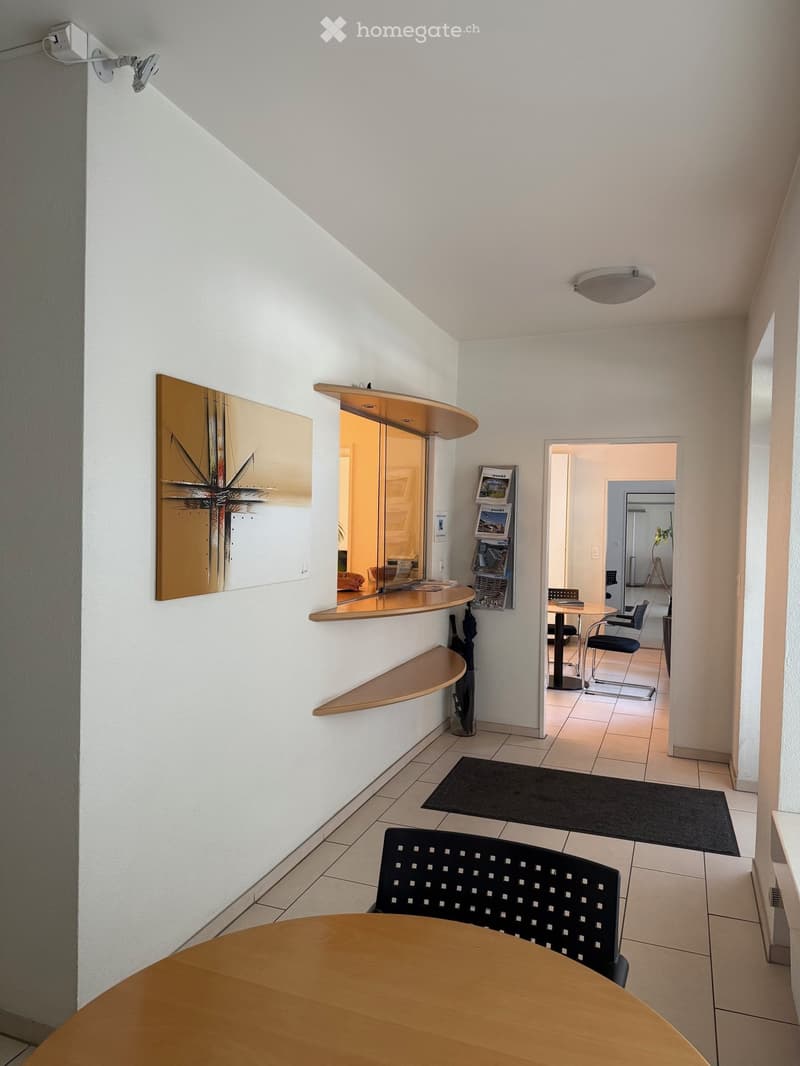 A modern hallway with a round wooden table, black chair, shelves, and a mirror on the wall. The area is well-lit with white walls and tiled flooring.