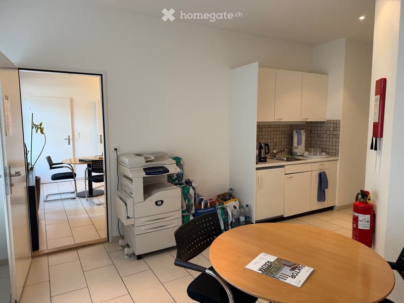 A furnished room with a small kitchen area, a table with chairs, a printer, and various items including bottles and a fire extinguisher.
