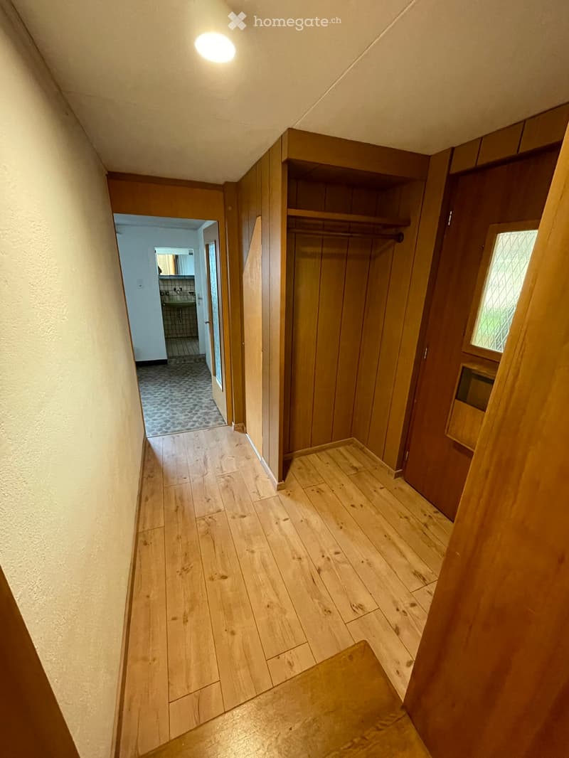 Wooden hallway with light wooden flooring, built-in wooden closet with shelves, and a small window on the right side. The hallway leads to a bathroom with a sink and tiled walls visible in the background.