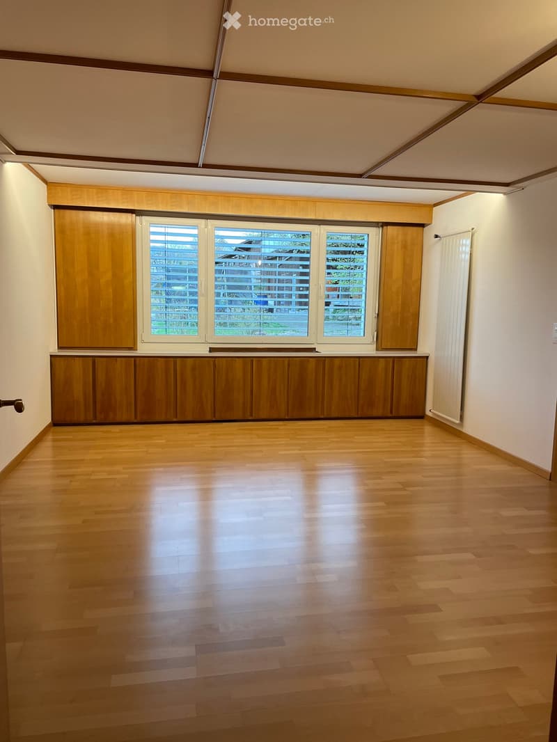 Empty room with wooden flooring, large windows with blinds, and built-in wooden cabinetry along one wall.