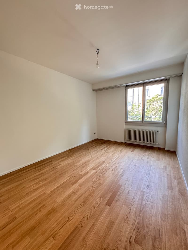 Empty room with wooden flooring, white walls, a single window with a radiator below, and a hanging light fixture.