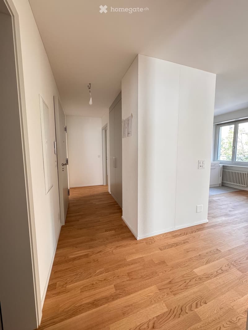 Empty hallway with wooden flooring, white walls, and several doors leading to other rooms. Natural light comes from a window at the end of the hallway.