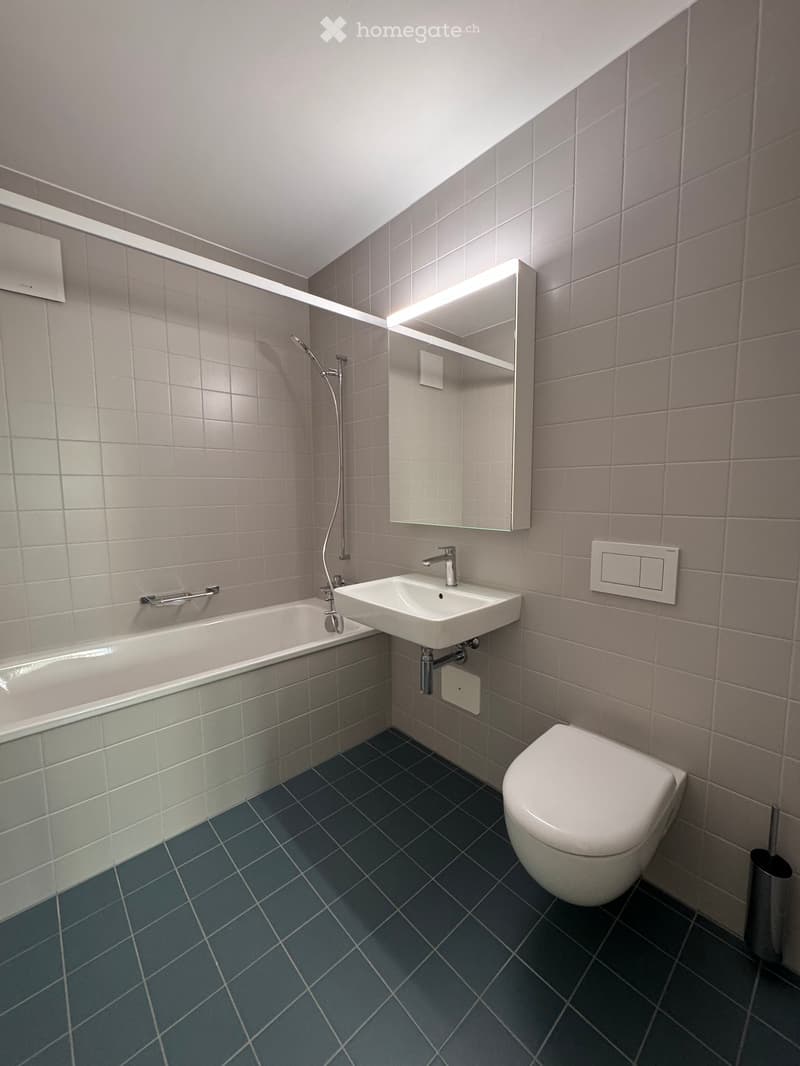 Modern bathroom with grey tiled walls, dark blue tiled floor, bathtub, sink, toilet, and illuminated mirror.
