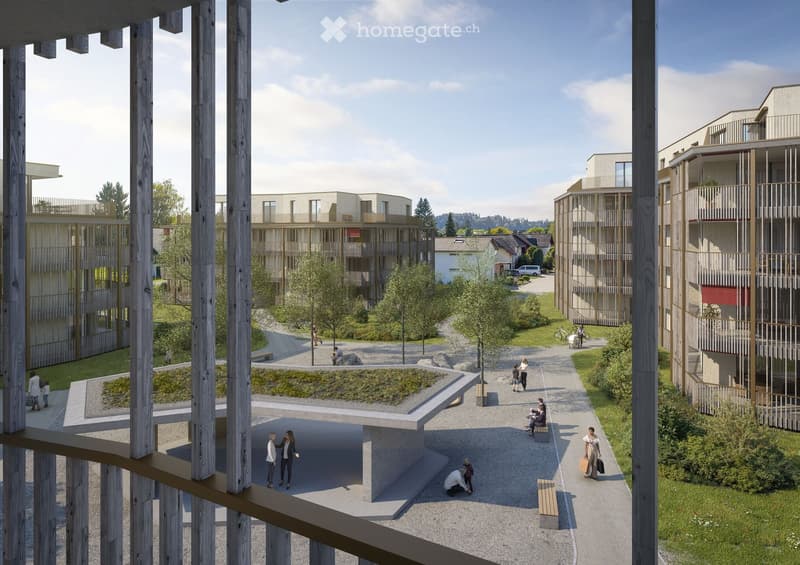 A modern residential complex with multiple buildings, green spaces, pathways, and people walking around. The area includes raised ground floors, benches, and landscaped gardens.
