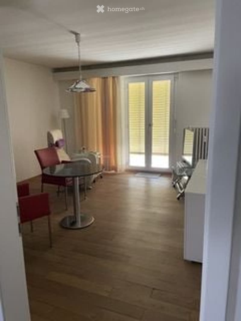 Bright living room with wooden flooring, large glass doors leading to a balcony, a round dining table with chairs, a lamp, and a TV screen.