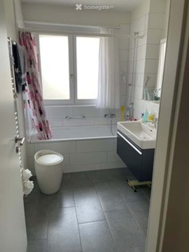 Modern bathroom with a bathtub, toilet, sink, and a window with a curtain. The floor is tiled in dark gray, and the walls are white.