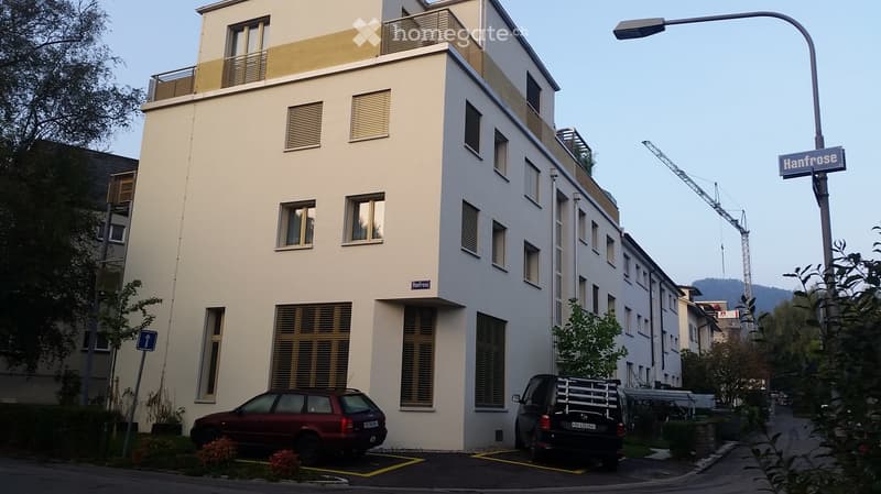 Modern multi-story building with white facade, large windows, and balconies. Two cars are parked in front, and there is a street sign reading 'Hanfrose'.
